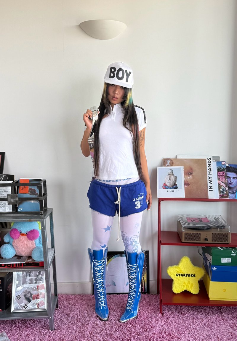Young woman with long hair wearing a white shirt, blue shorts, white "BOY" cap, patterned tights, and blue lace-up boots standing indoors.