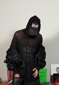 Sudadera de malla negra con un corte oversize, que presenta mangas de satén negro y un logotipo impreso. Se lleva con una gorra negra desgastada.