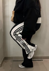 Sudadera negra con capucha, camiseta blanca de estilo en capas, pantalones deportivos Adidas en negro y blanco con tres rayas, zapatillas plateadas, sosteniendo una pequeña bolsa marrón claro.