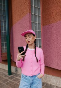Roza puhasta cardigan z gumbi, v kombinaciji s svetlo modrimi jeans hlačami. Roza kapa nošena nazaj, drži pametni telefon. Ozadje z teksturirano steno.