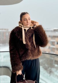 Veste courte en fausse fourrure marron à manches longues, avec un pull à capuche beige clair en dessous et une finition texturée. Pantalon foncé et sac à main.