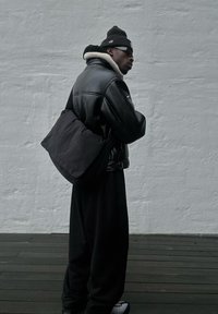 Giacca di pelle nera con colletto in shearling, pantaloni oversize neri e una borsa a tracolla nera. Sneakers grigie e un berretto nero.