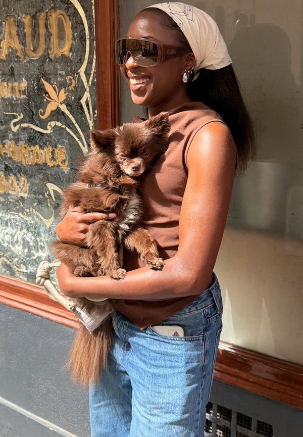 Hymyilevä nainen aurinkolaseissa ja päähuivissa pitää pientä ruskeaa koiraa, seisoo ulkona vintage-kaupan ikkunan lähellä.