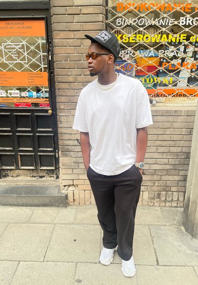 Camiseta blanca de cuello redondo, pantalones negros de corte holgado, reloj plateado, zapatillas blancas, gorra negra, gafas de sol tintadas; de pie contra una pared de ladrillo.