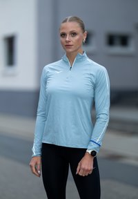 Svijetloplavi dugi sportski top s patentnim zatvaračem, s bijelim detaljima i teksturiranom tkaninom. Uparen s crnim tajicama i pametnom narukvicom.