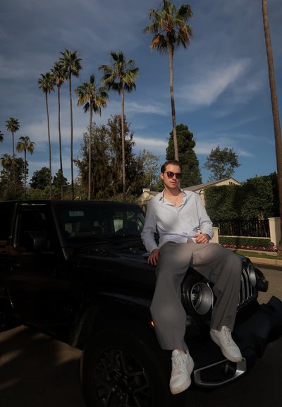 Joven con gafas de sol y camisa clara sentado sobre el capó de un SUV negro bajo altas palmeras en un día soleado en un barrio suburbano.