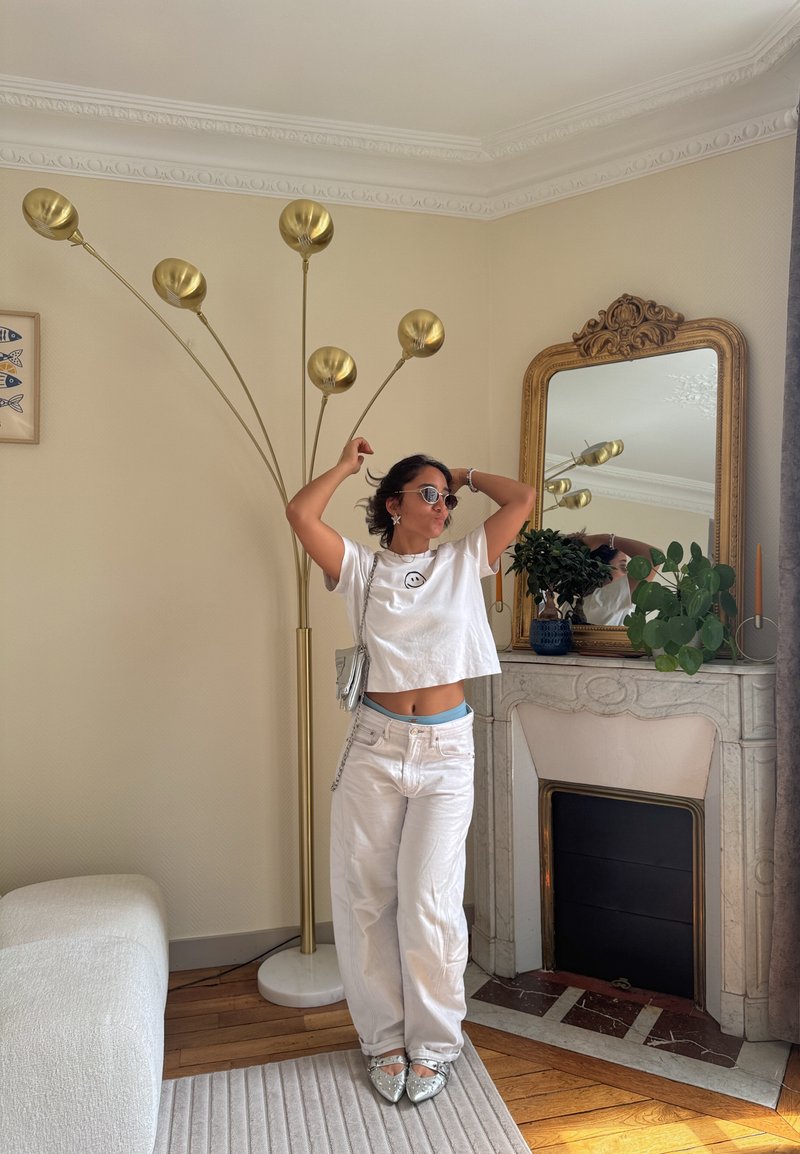 Femme vêtue de blanc et portant des lunettes de soleil pose avec les bras levés près d'une lampe sur pied dorée et une cheminée décorée avec un miroir et des plantes.