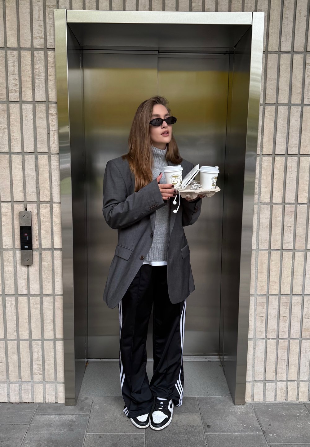 Eine Person hält ein Tablett mit zwei Kaffeetassen, trägt einen großen grauen Blazer, einen grauen Rollkragenpullover, schwarze Jogginghosen mit weißen Streifen und eine Sonnenbrille.