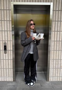 Eine Person hält ein Tablett mit zwei Kaffeetassen, trägt einen großen grauen Blazer, einen grauen Rollkragenpullover, schwarze Jogginghosen mit weißen Streifen und eine Sonnenbrille.