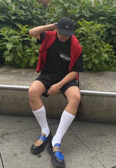 Camiseta negra con logo, suéter rojo drapeado sobre los hombros, pantalones cortos negros, calcetas blancas hasta la rodilla y zapatos negros con acentos azules y marrones.