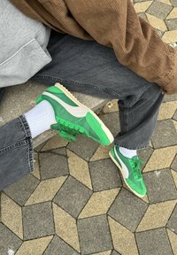 Zapatillas verdes de ante y malla, con una franja blanca, cordones verdes y una suela beige. Llevadas con jeans grises y calcetines blancos.