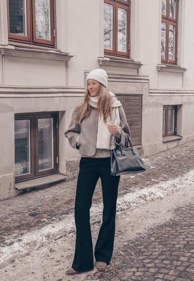 Mujer con ropa de invierno, gorro blanco y bufanda, llevando un bolso negro en una calle adoquinada nevada junto a un edificio beige con ventanas.