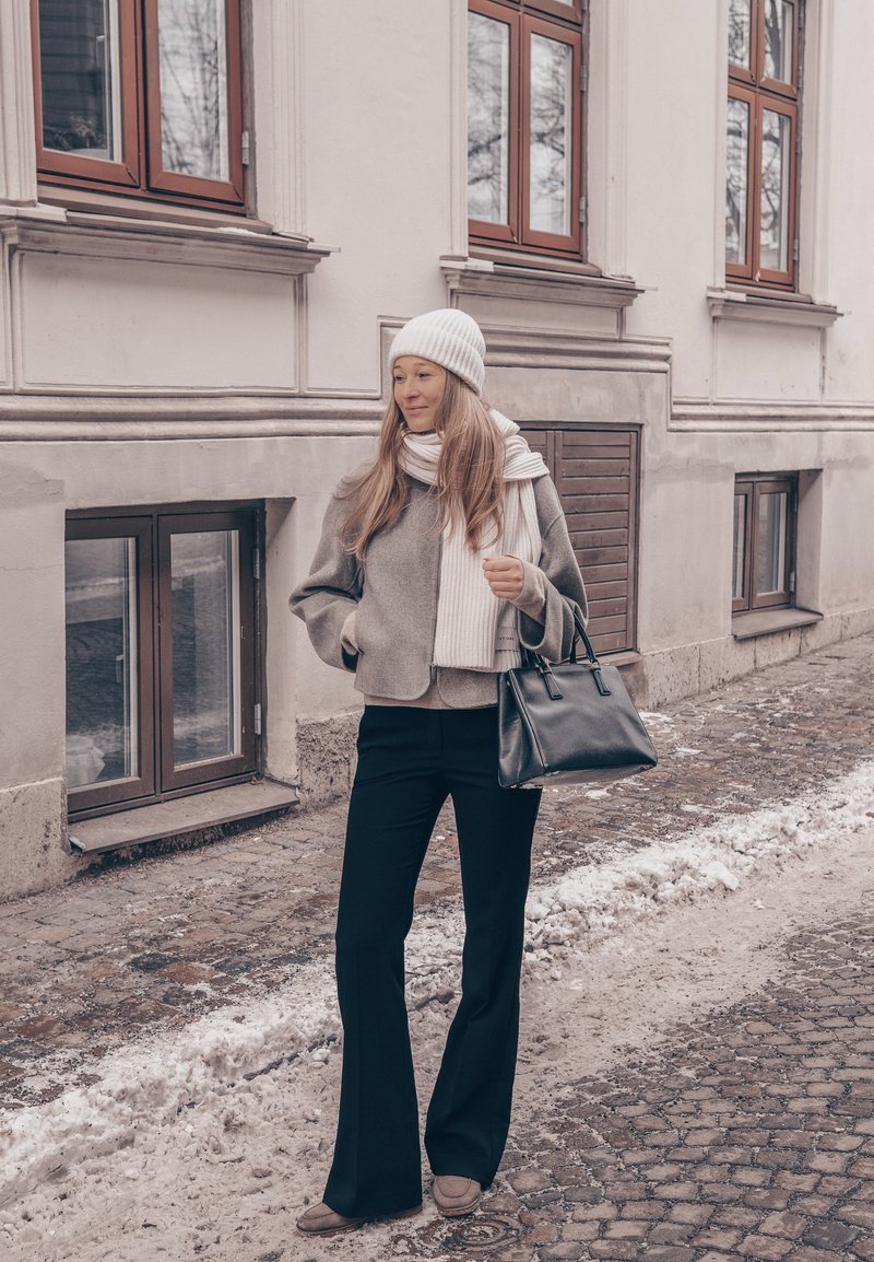 Femme en vêtements d'hiver avec un bonnet blanc et une écharpe, portant un sac à main noir dans une rue pavée enneigée près d'un bâtiment beige avec des fenêtres.