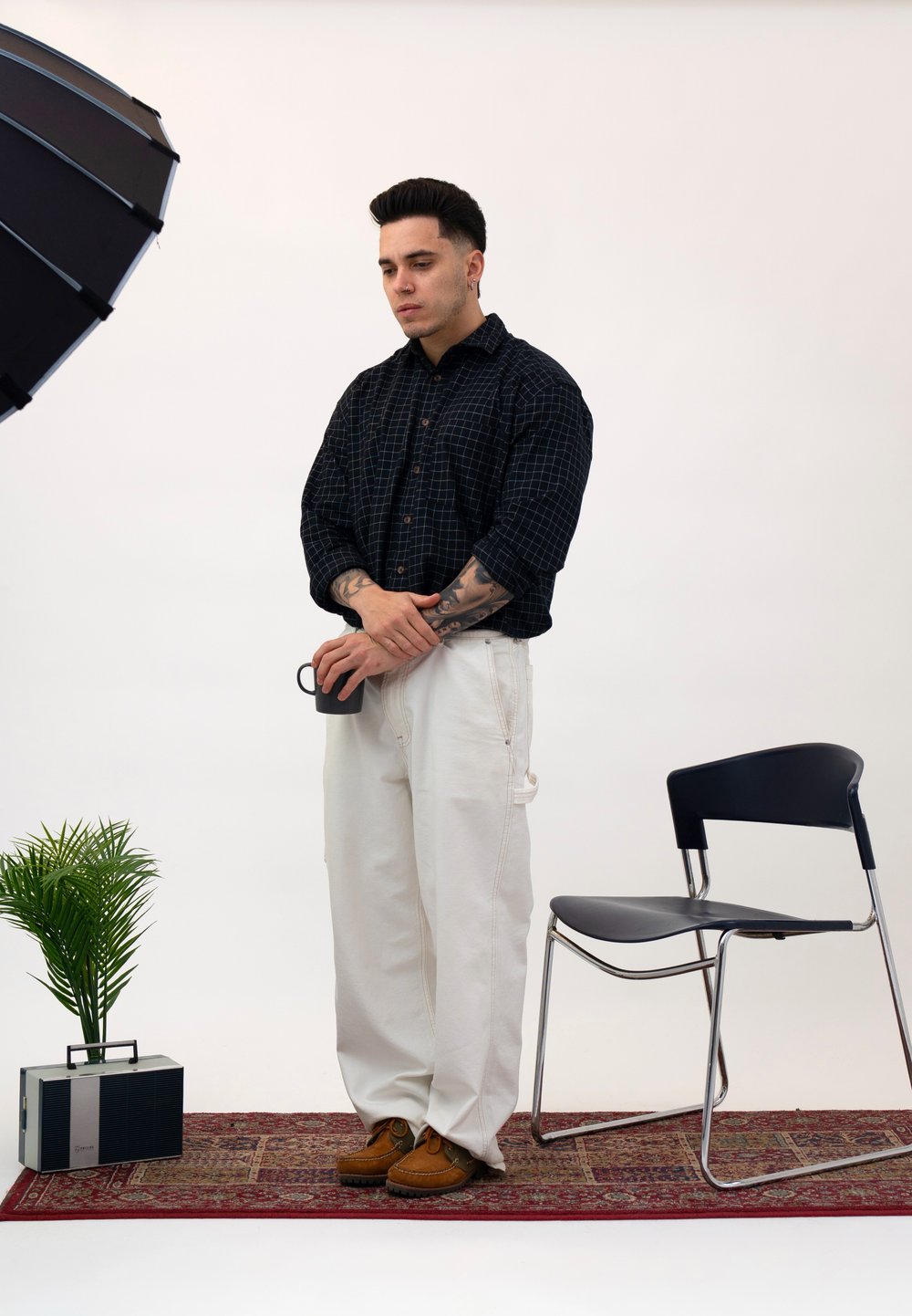 Mannequin portant une chemise noire à carreaux, un pantalon blanc et des chaussures marron, tenant une tasse. Une chaise pliante noire et une plante en pot sont à proximité sur un tapis.
