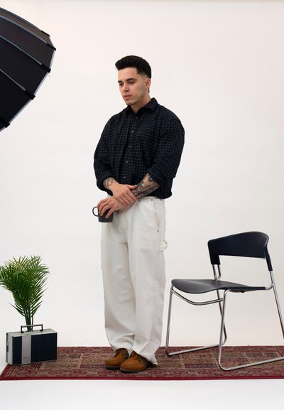 Modelo con camisa negra a cuadros, pantalones blancos y zapatos marrones, sosteniendo una taza. Cerca, una silla negra plegable y una planta en maceta sobre una alfombra.