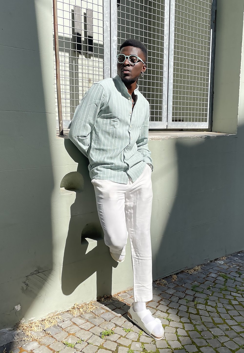 Lichtgroene gestreepte shirt, witte broeken en witte sandalen. Leunend tegen een muur naast een raam met een rasterpatroon. Zonovergoten achtergrond.