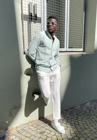 Lichtgroene gestreepte shirt, witte broeken en witte sandalen. Leunend tegen een muur naast een raam met een rasterpatroon. Zonovergoten achtergrond.