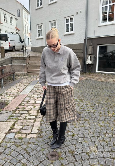 Joven con sudadera gris, pantalones cortos largos a cuadros, botas negras y gafas, de pie en una calle empedrada cerca de escaleras y edificios.