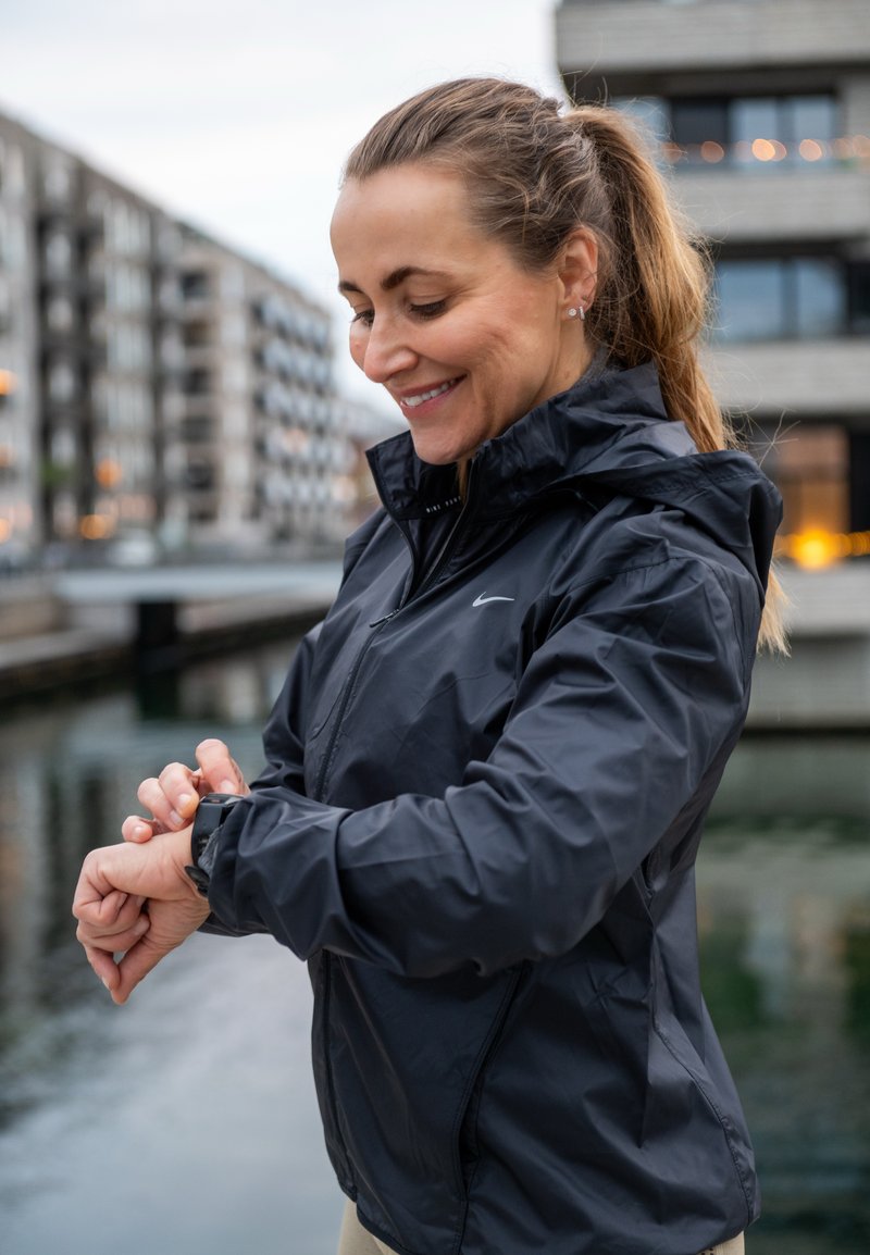 Jachetă neagră, ușoară, cu fermoar frontal și glugă, purtată peste un top deschis la culoare. Persoana poartă un ceas la încheietura mâinii stângi.