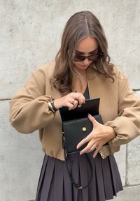 Chaqueta bomber marrón, billetera de cuero negra con detalles en dorado y falda negra con pliegues. La persona parece estar sacando artículos de la billetera.