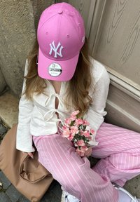 Gorra rosa de los New York Yankees, parte superior blanca texturizada, pantalones a rayas rosas, sosteniendo flores rosas, con una bolsa beige al lado. Sentada contra una pared.