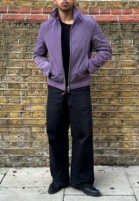 Violetti bomber-takki pystykkauluksella, vetoketjulla ja sivutaskuilla, yhdistettynä mustiin leveälahkeisiin housuihin ja mustiin kenkiin. Teksturoitu kangas.