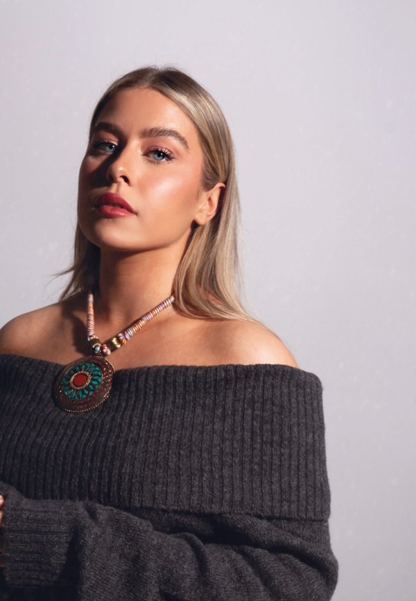 Halsband med en rund hängande detalj som har ett turkos- och röddesign, trädd på en pärlglittrande kedja med guldfinish. Modell bär en mörk tröja.