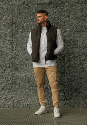 Chaleco acolchado marrón oscuro sobre una camisa blanca de manga larga, combinado con pantalones cargo beige y zapatillas blancas. De pie contra una pared texturizada gris.