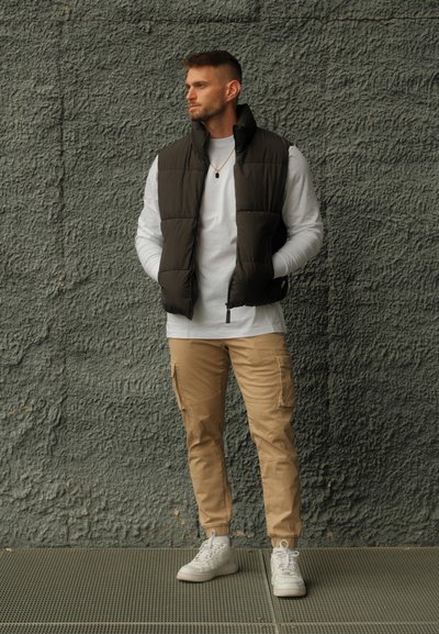 Chaleco acolchado marrón oscuro sobre una camisa blanca de manga larga, combinado con pantalones cargo beige y zapatillas blancas. De pie contra una pared texturizada gris.