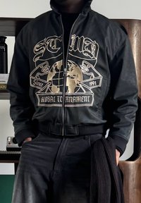 Personne portant une veste noire zippée avec un globe doré et le texte « SCMB Global Tournament Since 1991 », tenant une écharpe en tricot noir dans une main.
