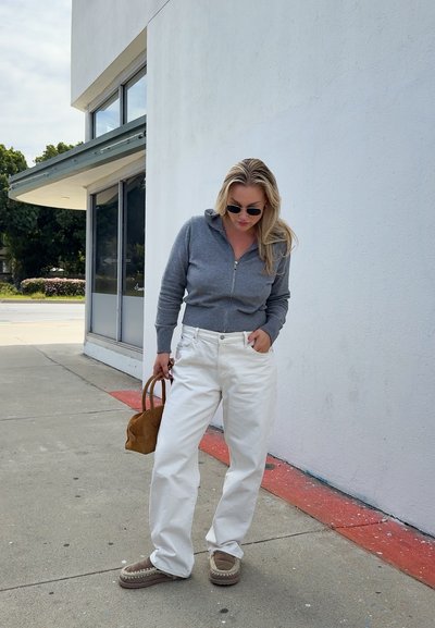 Mujer con suéter gris con cremallera, pantalones blancos, zapatos marrones y gafas de sol sostiene un bolso marrón mientras está de pie en la acera junto a la pared de un edificio blanco.