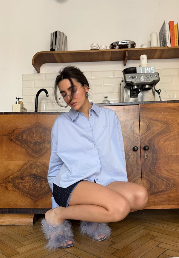 Young woman in an oversized blue shirt, black shorts, and fluffy slippers squats on a wooden floor in a kitchen with an espresso machine and wooden cupboards.