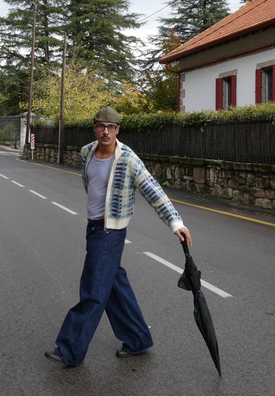 Hombre caminando por una calle, vestido con un cárdigan de punto con estampado en verde y azul, camiseta gris, vaqueros azules holgados y sosteniendo un paraguas negro.