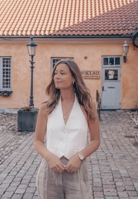 White sleeveless blouse with buttons, paired with light beige linen trousers. Outdoor cobblestone setting with an orange wall and window.