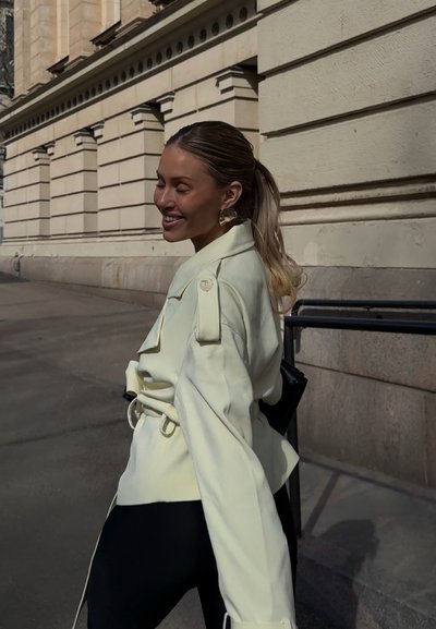 Mujer sonriendo con los ojos cerrados, lleva una chaqueta clara con cinturón y grandes pendientes, caminando junto a un edificio de piedra a la luz del día.