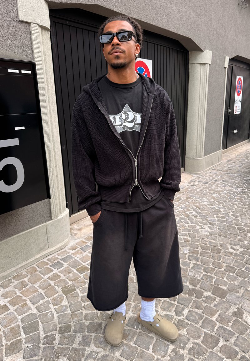 Homme portant un sweat à capuche noir, un t-shirt graphique et un long short noir se tient sur un trottoir pavé devant des portes de garage sombres.