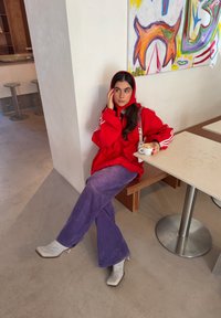 Γυναίκα με κόκκινο φούτερ με κουκούλα και μωβ παντελόνι κάθεται σε καφέ κρατώντας μια κούπα καφέ, ακουμπώντας σε λευκό τοίχο κάτω από πολύχρωμη αφηρημένη τέχνη.