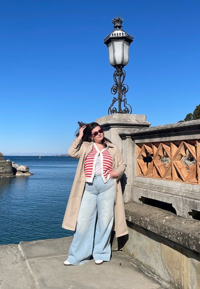 Un abrigo trench beige sobre un suéter de rayas rojas y blancas, jeans de pierna ancha azul claro y zapatos blancos, con un farol de metal y un fondo de waterfront.