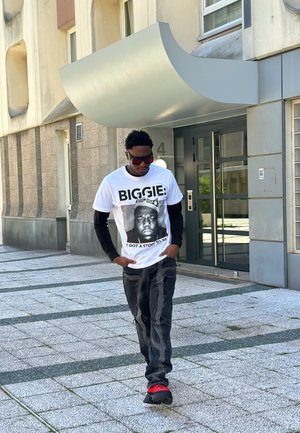 Camiseta blanca con un estampado gráfico de un hombre coronado, texto "BIGGIE: TENGO UNA HISTORIA QUE CONTAR", combinada con jeans negros desgastados y zapatillas rojas.