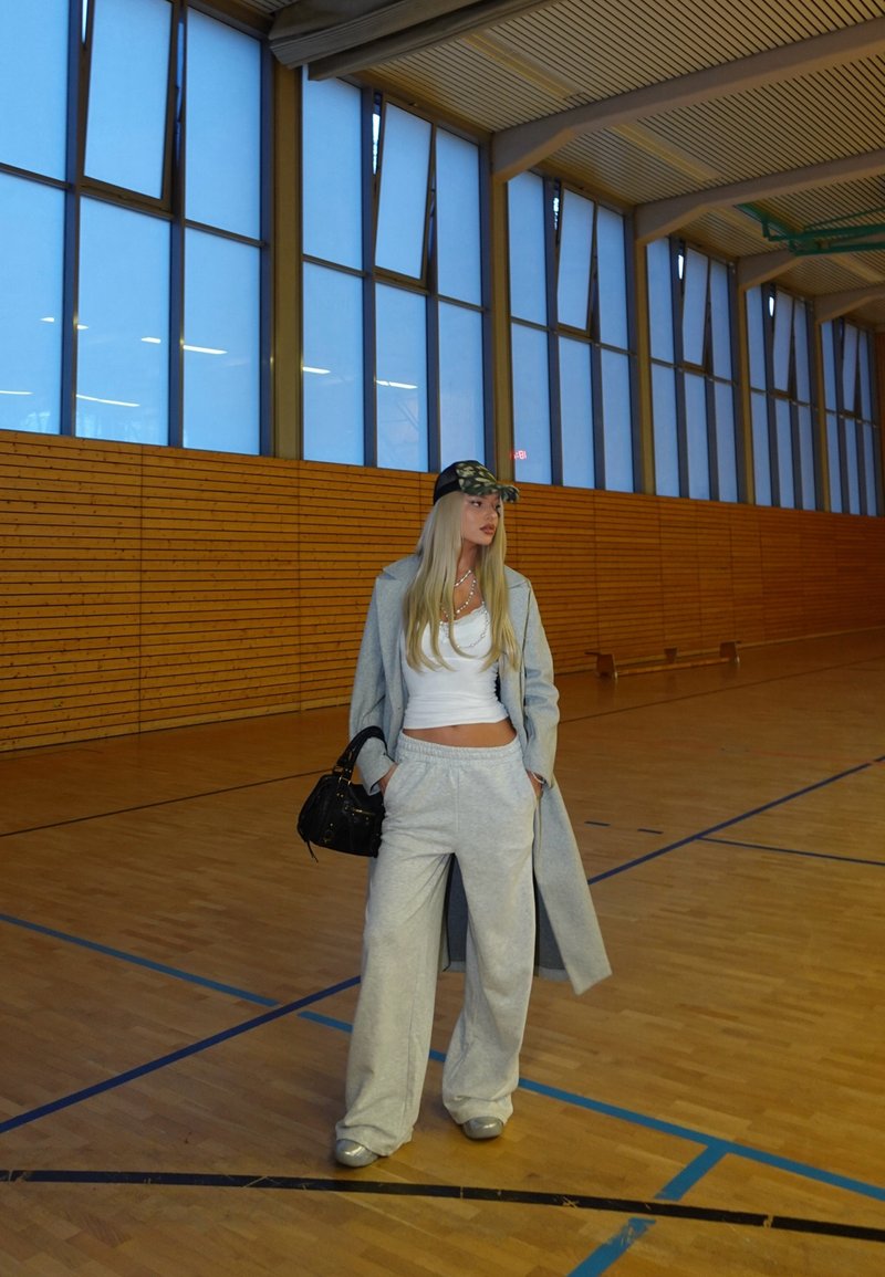 Γκρι φόρμες, λευκό crop top, μακρύ γκρι παλτό και καπέλο camo. Μαύρη τσάντα και ανοιχτό γκρι παπούτσια, σε γυμναστήριο με ξύλινα τοιχώματα και μεγάλα παράθυρα.