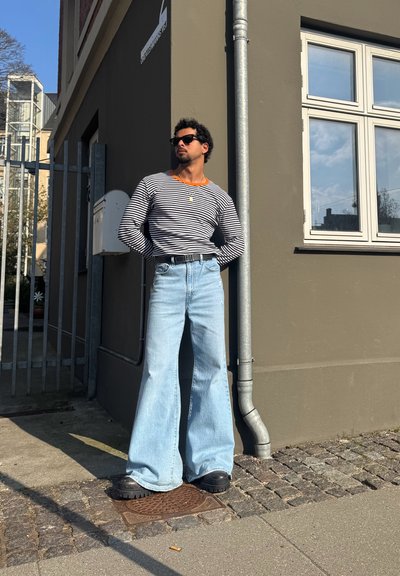 Camisa de rayas de manga larga con cuello naranja, jeans de pierna ancha azul claro, zapatos gruesos negros y gafas de sol, apoyado contra una pared.