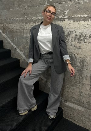 Blazer gris de gran tamaño con mangas enrolladas, combinado con pantalones rectos grises. Camiseta blanca debajo y zapatillas de tonos neutros. Fondo texturizado.
