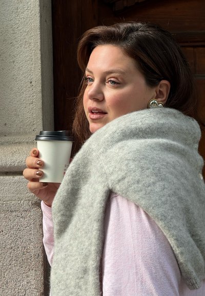 Bufanda de lana gris suspendida sobre una blusa de color rosa claro, con una mano sosteniendo una taza de café blanca con una tapa negra. Joyería en las orejas con forma de corazón.