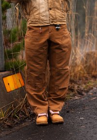 Personne portant un pantalon de travail marron et des bottes marron clair, debout sur un chemin asphalté mouillé près d'une clôture et d'herbe sèche.