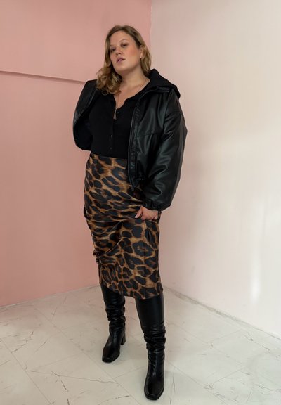 Mujer con chaqueta de cuero negra, camiseta negra, falda de leopardo y botas altas negras de rodilla, de pie sobre un suelo de mármol frente a una pared rosa y blanca.