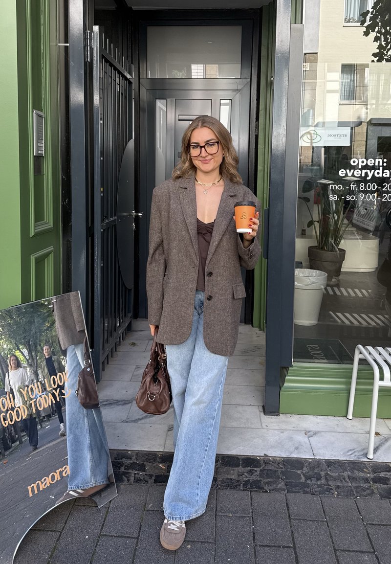 Rjav kariran oversized blazer, črna rebrasta majica, svetlo modri široki kavbojki, rjava torbica, drži skodelico kave, stoji blizu vrat.