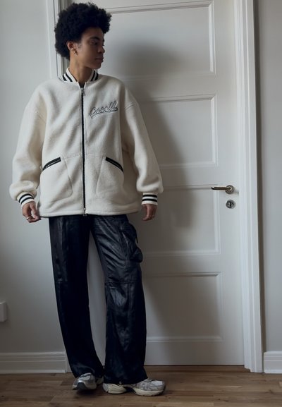 Chaqueta de forro polar blanca con cremalleras y ribetes negros, y logo bordado, combinada con pantalones cargo negros holgados y zapatillas grises.
