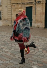 Kolorowy płaszcz z futerka sztucznego w mieszance niebieskiego, różowego i czerwonego. Para czarnych kozaków do kolan, czerwony szal w wzory oraz mała czerwona torebka z zawieszkami.