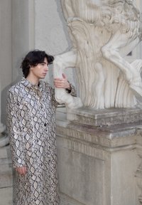 Jeune personne portant un manteau à motif peau de serpent, tenant la jambe d'une statue en marbre blanc dans un décor architectural en pierre ornée.