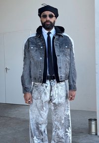 Sort og grå denimjakke med hvide accenter, sammen med hvide bukser med et tekstureret blomstermønster. Tilbehøret inkluderer en sort beret og runde solbriller.